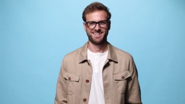 Genç adam gülümsüyor, kollarını kavuşturuyor, kameraya bakıyor ve mavi arka planda gözlük takıyor.