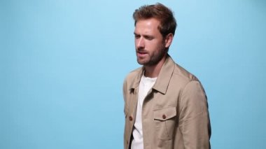 Havalı poz veren, bakışlarını çeviren, şehvetli bir şekilde yüzüne dokunan, kollarını kavuşturan ve mavi arka planda gülümseyen bir adam.