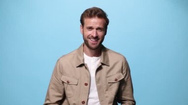 Mutlu sıradan bir adam gülüyor ve eğleniyor, poz veriyor, boynuna dokunuyor, ceketini tamir ediyor, yana dönüyor ve mavi arka plandaki kameraya bakıyor.