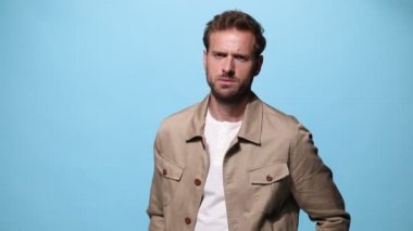 Yakışıklı, sıradan bir adam başka tarafa bakıyor, poz veriyor ve bir tarafa dönüyor, ceketini tamir ediyor ve mavi arka plandaki kameraya bakıyor.