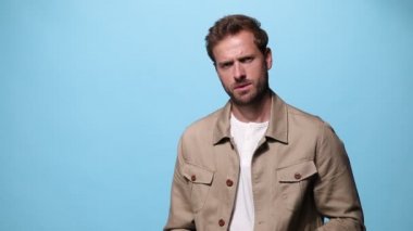 Moda adamı ceketini düzeltiyor, kollarını kavuşturuyor, yavaşça yüzüne dokunuyor ve mavi arka plandaki kameraya bakıyor.