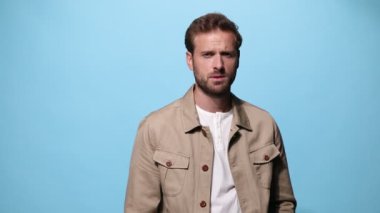 Seksi moda adamı kameranın derinliklerine bakıyor, poz veriyor, yavaşça bir kolunu katlıyor, çenesine dokunuyor, ceketini mavi arka plana yerleştiriyor.