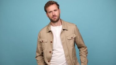 Genç manken ellerini cebine sokuyor, gülümsüyor ve yana dönüyor, kameraya bakıyor, kollarını kavuşturuyor ve mavi arka plana bakıyor.