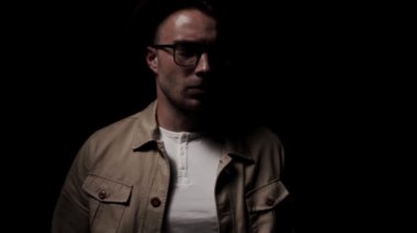 Karanlıktan gelen kendine güvenen genç adam, ceketini düzeltiyor, şehvetli bir şekilde yüzüne dokunuyor, yukarı bakıyor, şapka ve gözlük takıyor, siyah arka planda bir gölgede yürüyor.