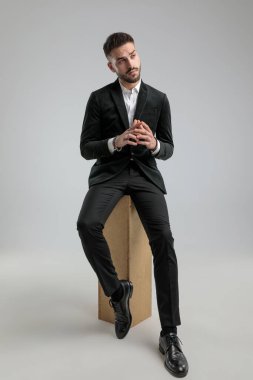 Genç işadamı bakışlarını başka yöne çeviriyor, ellerini birarada tutuyor ve gri arka planda oturuyor.