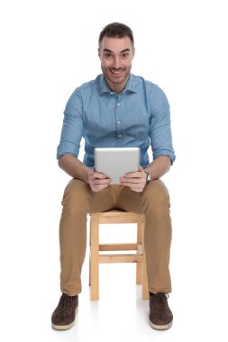 Beyaz stüdyo arka planında bir sandalyede otururken gülümseyen ve tablet tutan zeki bir adam.