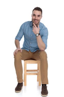 Mutlu, zeki, sıradan bir adam, çenesine sarılıp düşünüyor, gülümsüyor ve beyaz stüdyo arka planında bir sandalyede oturuyor.