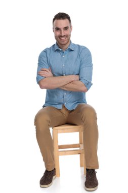 Beyaz stüdyo arka planında bir sandalyede otururken elleri çaprazlanmış gülümseyen zeki bir adam.