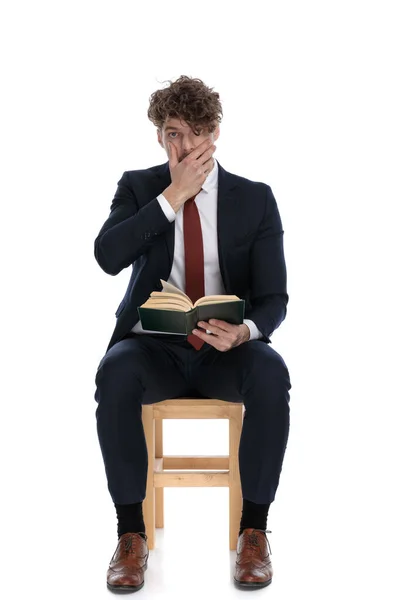 聪明而随和的生意人捂着嘴 拿着书 靠着白色的背景坐着 — 图库照片