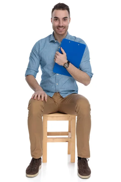 幸せなスマートカジュアル男保持クリップボードと笑顔上の椅子に座っている間にホワイトスタジオの背景 — ストック写真