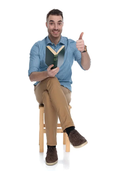 ポジティブなスマートカジュアル男保持本と与え親指上の椅子に座っている間にホワイトスタジオの背景 — ストック写真