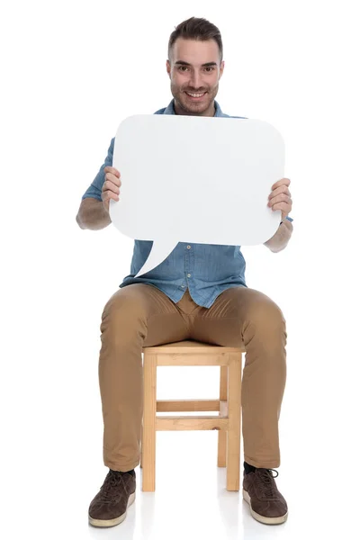 積極的なスマートカジュアルな男笑顔と空のスピーチバブルを保持しながら 上の椅子に座っていますホワイトスタジオの背景 — ストック写真