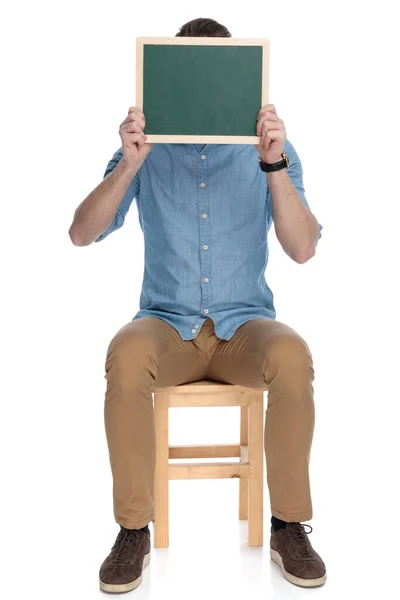 年轻聪明的休闲男子坐在白色画室背景的椅子上 用黑板盖住脸 — 图库照片