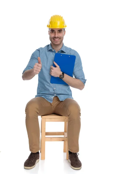 ハッピースマートカジュアル男保持クリップボード ジェスチャーOkと身に着けています安全ヘルメットながら 上の椅子に座って白いスタジオの背景 — ストック写真