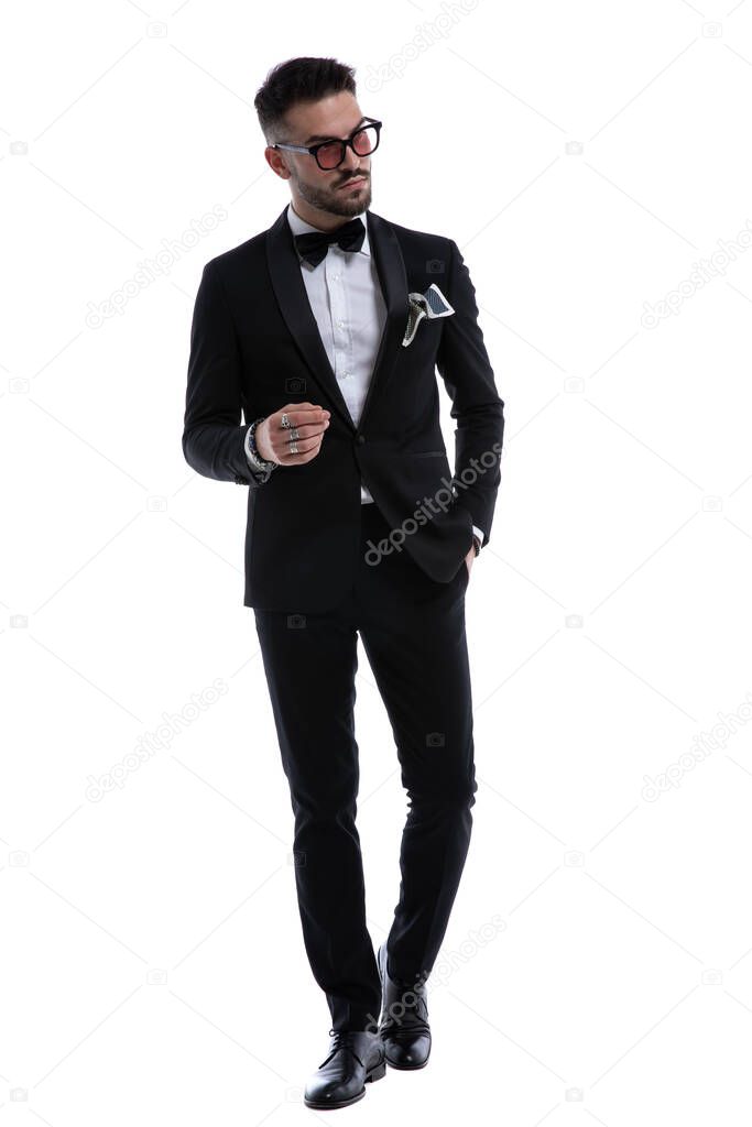 elegant young guy in tuxedo holding hands in a fashion pose looking to side and walking isolated on white background