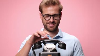 Mavi polo tişörtlü mutlu genç adam gözlük takıyor, eski bir telefonu kaldırıyor, kaşlarını çatıyor, tartışıyor ve bağırıyor, pembe arka planda takılıyor.