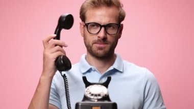 Mavi polo tişörtlü gülümseyen, gözlüklü, eski bir telefona cevap veren, kızgın bir yüz ifadesi takınan, şakağı işaret eden, bağıran ve yumruk hareketi yapan, pembe arka planda asılı duran