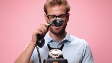 Mavi polo tişörtlü mutlu genç adam eski bir telefonu cevaplıyor, şok olmuş bir ifade takınıyor, yumruklarını havaya kaldırıyor ve zaferi kutluyor, pembe arka planda gülüyor ve çığlık atıyor.