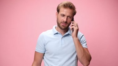 Mavi polo tişörtlü sıradan bir genç telefonda konuşuyor, harika sohbet ediyor ve gülümsüyor, telefonu arka cebine koyuyor pembe arka planda.