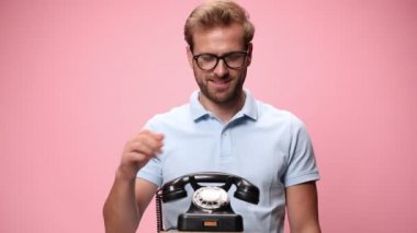 Mavi polo tişörtlü, gözlüklü genç bir adam, eski telefondan konuşuyor, gülüyor, gümüş kupa tutuyor ve pembe arka planda kutlama yapıyor.