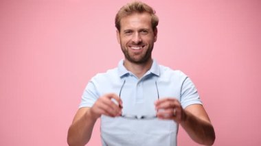 Mavi polo tişörtlü mutlu genç adam gözlük takıyor, gülümsüyor, şaşırmış bir yüz ifadesi takınıyor, kollarını kavuşturup pembe arka planda başını sallıyor.
