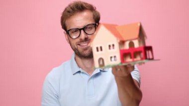 Mavi polo tişörtlü sıradan bir genç ev modelini tavsiye ediyor, gülümsüyor, yumruklarını havaya kaldırıyor ve kutluyor, pembe arka planda gülüyor ve çığlık atıyor.