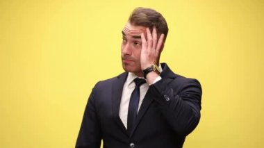 Yakışıklı işadamı yana bakıyor, kafasını kaşıyor, sonra harika bir fikir aklına geliyor ve sarı arka plana işaret ederken mutlu bir şekilde şaşırıyor.