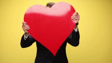 Yakışıklı iş adamı büyük kırmızı kalbini kameraya gösteriyor, sarı arka planda neşeli.