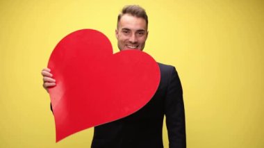 Seksi iş adamı büyük kırmızı bir kalp sunar sonra onu, kalbini ve sarı arka plandaki kamerayı gösterir.