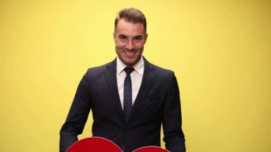 Çekici iş adamı yavaşça büyük kırmızı bir kalp kaldırıyor sonra da onu sarı arka planda kameraya gösteriyor.