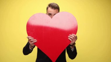 Genç ve çekici bir iş adamı büyük kırmızı bir kalbin arkasına saklanıyor. Sonra üzgün yüzünü gösteriyor ve kalbini sarı arka planda kollarında tutuyor.