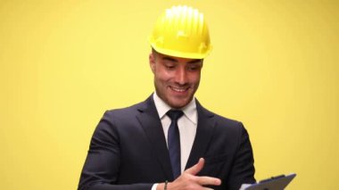 Sarı kask takan genç ve seksi iş adamı önce dosyasına sonra da kameraya bakıyor ve sarı arka planda yumruğunu kaldırıyor.