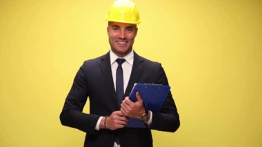 Genç çekici iş adamı kask takıyor, elinde bir pano tutuyor, tek koluyla tutuyor, sarı arka plandaki kameraya panoyu gösteriyor.