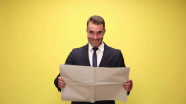 Çekici iş adamı gazeteyi tutuyor ve yavaşça kaldırıyor, arkasına saklanıyor ve sarı arka plandaki kameraya gösteriyor.