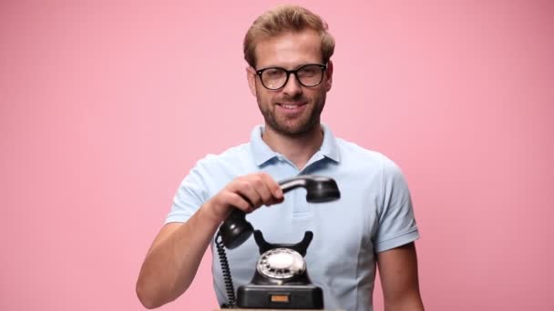 穿着蓝色马球衫的随意微笑的家伙拿起旧的旋转电话 展示房子模型 并在粉色背景上打盹 — 图库视频影像