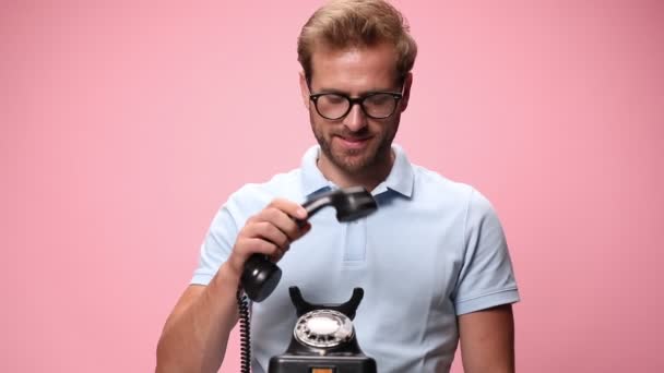 穿着蓝色马球衫 戴着眼镜的年轻而随意的小伙子笑着 一边接电话 一边用粉红的背景做手势 — 图库视频影像