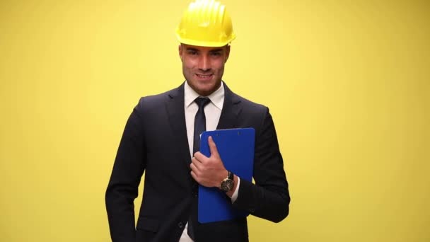 英俊的商人戴着头盔 拿着剪贴板 一只手插在口袋里 望着旁边 微笑着 在黄色的背景上摆出姿势 — 图库视频影像