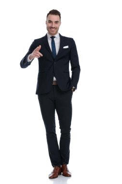 Kendine güvenen işadamı, beyaz stüdyo arka planında dururken elini cebine atarak gülümsüyor ve ileri işaret ediyor.