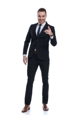 Kendine güvenen işadamı gülümseyip öne doğru işaret ederken beyaz stüdyonun arka planında duruyor.