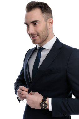 Mutlu iş adamı ceketini düzeltiyor ve beyaz stüdyo arkasında dururken gülümsüyor.