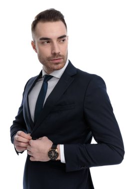 Beyaz stüdyo arka planında dikilirken ceketi ayarlayan ve omzunun üstünden bakan çekici bir iş adamı.