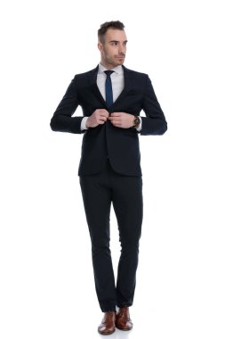 Ciddi bir işadamı, beyaz stüdyo arka planında yürürken ceketini düzeltip başka tarafa bakıyor.