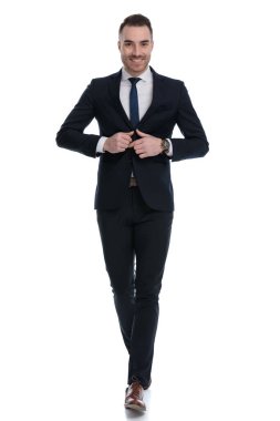 Mutlu iş adamı beyaz stüdyo arka planında yürürken ceketini düzeltiyor ve gülümsüyor.