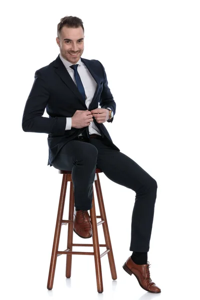 積極的なビジネスマンは笑ってジャケットを調整しながら 白いスタジオの背景にあるスツールに座って — ストック写真