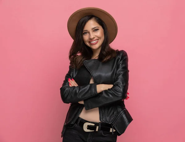 Cheerful fashion model smiling with her hands folded, wearing hat while standing on pink studio background