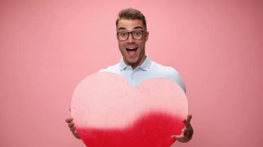 Model büyük kırmızı bir kalp tutuyor, heyecanlanıyor ve kalbini pembe arka planda kameraya veriyor.