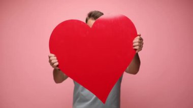 Genç ve çekici bir adam büyük kırmızı kalbini kameraya gösteriyor ve yavaşça pembe arka planda bırakıyor.