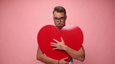 Çekici, genç, sıradan bir adam büyük kırmızı bir kalbe sarılıyor ve pembe arka planda üzgün ve huysuz.