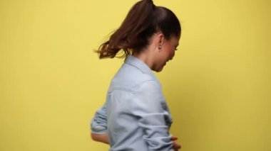 Mavi kot gömlekli hevesli genç bir kadın piruet yapıyor, gülümsüyor, kollarını havaya açıyor ve sarı arka planda dans ediyor.