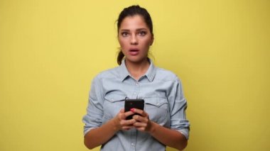 Şok olmuş bir kadın mavi kot gömlek giyiyor, kötü haberler okuyor, ağzı açık, elini alnında tutuyor, elini sarı arka planda tutuyor.